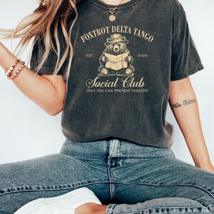 Puede incluir: Una camiseta gris oscuro con un gr&aacute;fico de estilo vintage de un oso con un sombrero leyendo un mapa. El texto "Fox Trot Delta Tango Est 2025 Social Club Only You Can Prevent Fascism" est&aacute; impreso en la camiseta.