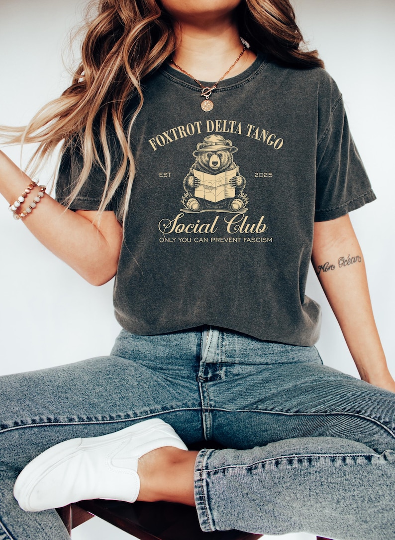 Puede incluir: Una camiseta gris oscuro con un gr&aacute;fico de estilo vintage de un oso con un sombrero leyendo un mapa. El texto "Fox Trot Delta Tango Est 2025 Social Club Only You Can Prevent Fascism" est&aacute; impreso en la camiseta.