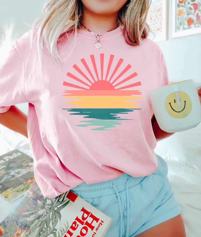 May include: A pink t-shirt with a retro sunset graphic featuring a red sun, yellow, orange, and blue stripes, and a teal and green reflection in the water.
