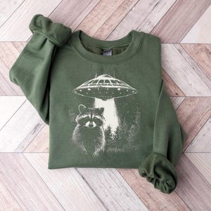 May include: Green sweatshirt featuring a graphic of a raccoon, a UFO, and a forest scene. The design is in white against the green fabric. The sweatshirt has long sleeves and a crew neck.