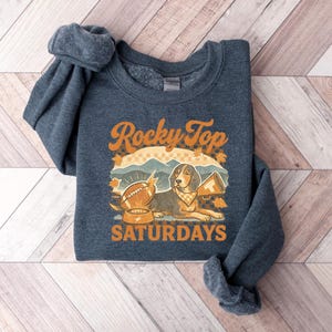 May include: A navy blue sweatshirt with the words "Rocky Top Saturdays" in orange. The graphic features a beagle with a bandana, a football, and a megaphone. The sweatshirt is folded on a wooden surface.