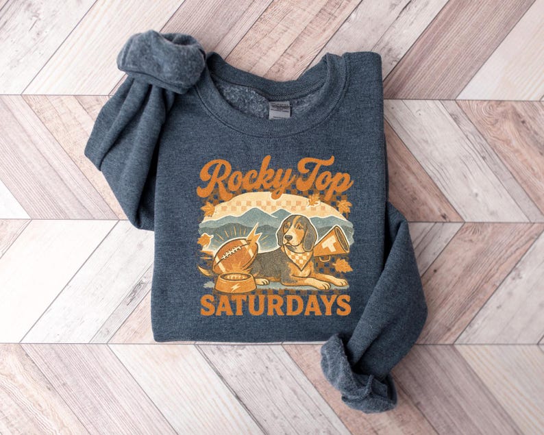 May include: A navy blue sweatshirt with the words "Rocky Top Saturdays" in orange. The graphic features a beagle with a bandana, a football, and a megaphone. The sweatshirt is folded on a wooden surface.