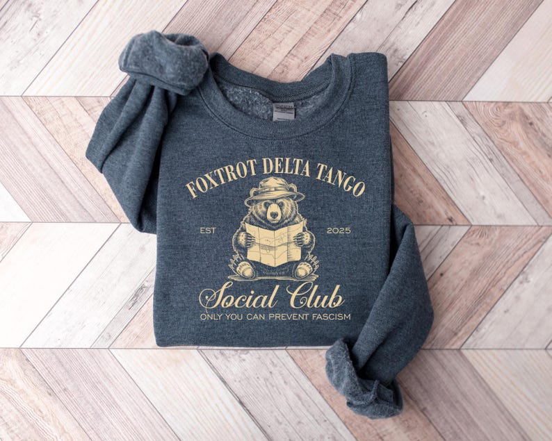 Puede incluir: Una sudadera gris oscuro con un gr&aacute;fico de un oso con un sombrero de guardabosques y sosteniendo un mapa. El texto "Foxtrot Delta Tango Social Club Est 2025 Only You Can Prevent Fascism" est&aacute; impreso en la sudadera.
