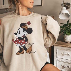 May include: Beige sweatshirt featuring a Minnie Mouse graphic. The cartoon character is depicted in a red polka-dot dress, brown shoes, and a hat with a flower. The Disney logo is printed on the lower right side of the graphic.
