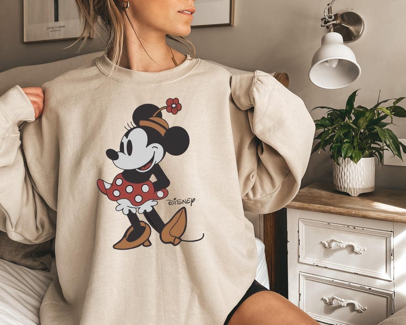 May include: Beige sweatshirt featuring a Minnie Mouse graphic. The cartoon character is depicted in a red polka-dot dress, brown shoes, and a hat with a flower. The Disney logo is printed on the lower right side of the graphic.