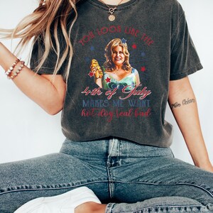 May include: A dark gray t-shirt with a distressed look featuring a woman holding a hot dog and the text "You look like the 4th of July makes me want hot dog real bad."