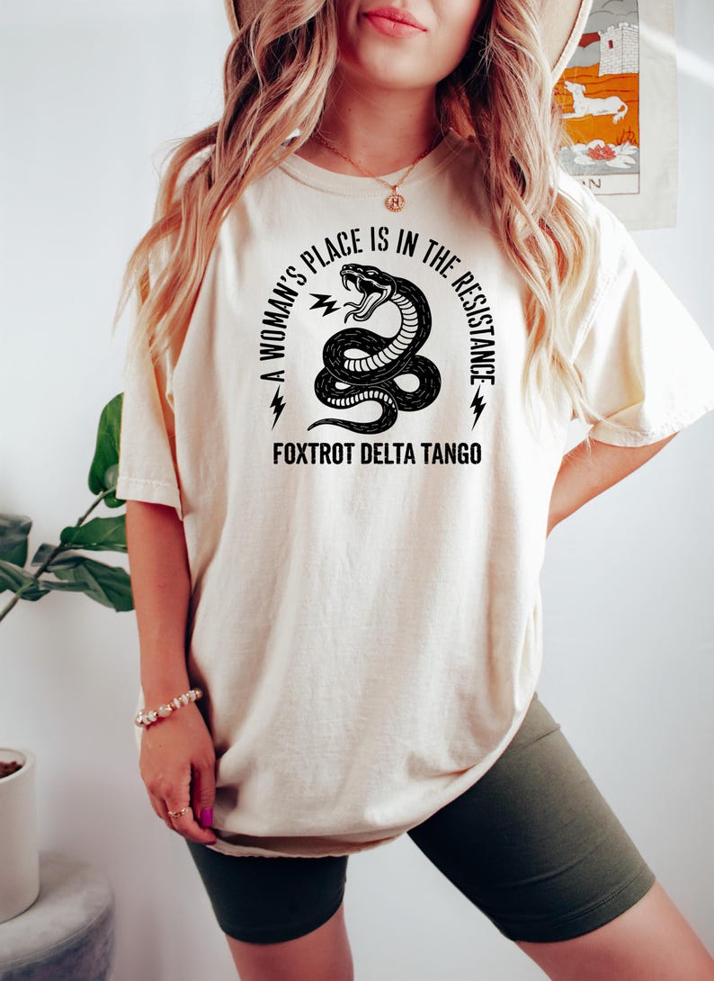 May include: A white t-shirt with a black graphic of a snake with lightning bolts and the text "A woman's place is in the resistance. Fox Trot Delta Tango".