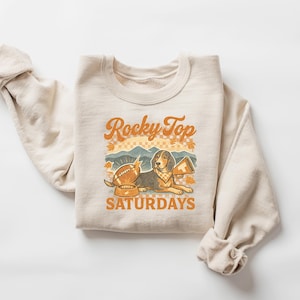May include: Cream-colored sweatshirt with the text "Rocky Top Saturdays" in orange and brown. The graphic features a beagle with a football, a megaphone, and a bowl. The design has a vintage, collegiate aesthetic.