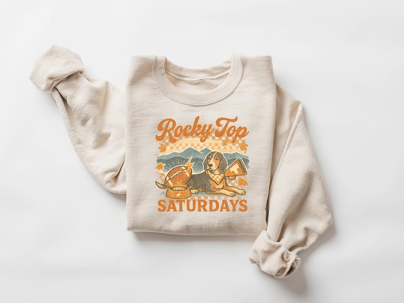 May include: Cream-colored sweatshirt with the text "Rocky Top Saturdays" in orange and brown. The graphic features a beagle with a football, a megaphone, and a bowl. The design has a vintage, collegiate aesthetic.