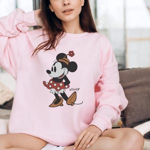 May include: A light pink sweatshirt featuring a Minnie Mouse graphic. The cartoon character is depicted in a red polka-dot dress, brown shoes, and a matching hat. The Disney logo is printed below the character.