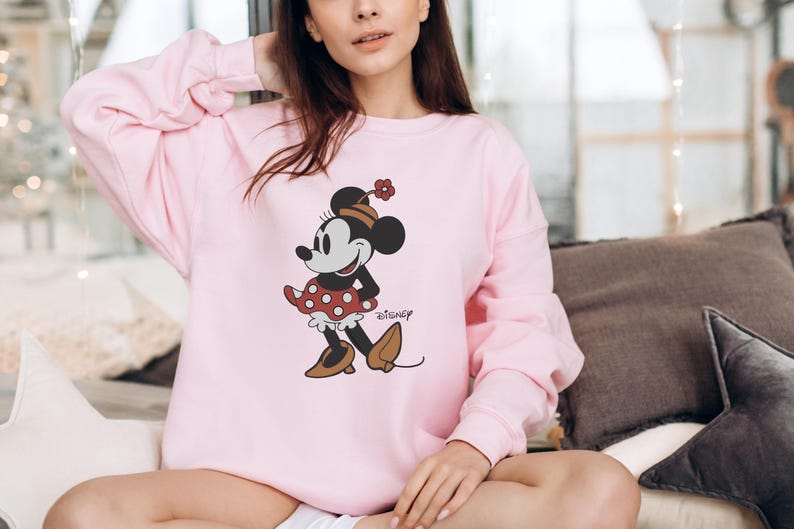 May include: A light pink sweatshirt featuring a Minnie Mouse graphic. The cartoon character is depicted in a red polka-dot dress, brown shoes, and a matching hat. The Disney logo is printed below the character.