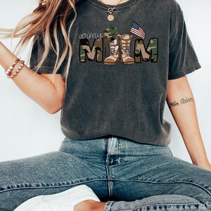 May include: A dark gray t-shirt with a camouflage print and the text "army mom" in white. The text is surrounded by a camouflage pattern and includes a military hat, boots, and an American flag.