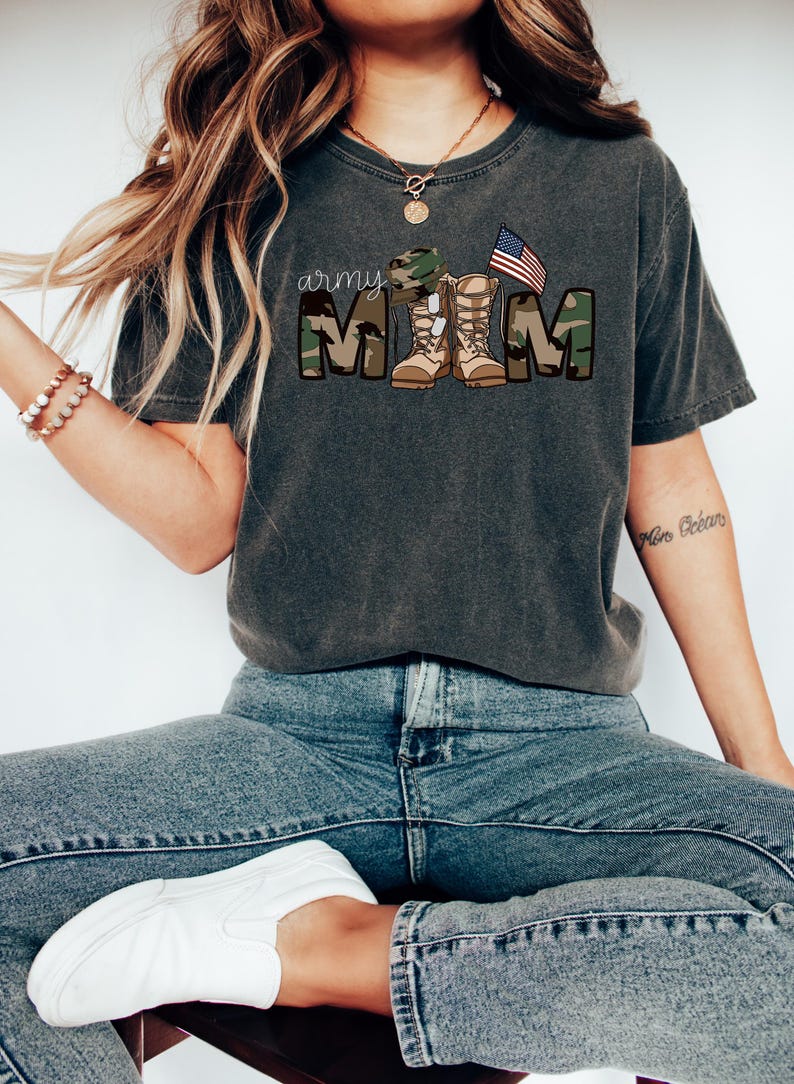 May include: A dark gray t-shirt with a camouflage print and the text "army mom" in white. The text is surrounded by a camouflage pattern and includes a military hat, boots, and an American flag.