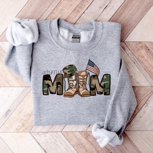 May include: Gray sweatshirt with a camouflage print that says "army mom" with a pair of boots and a military hat. There is an American flag above the boots.