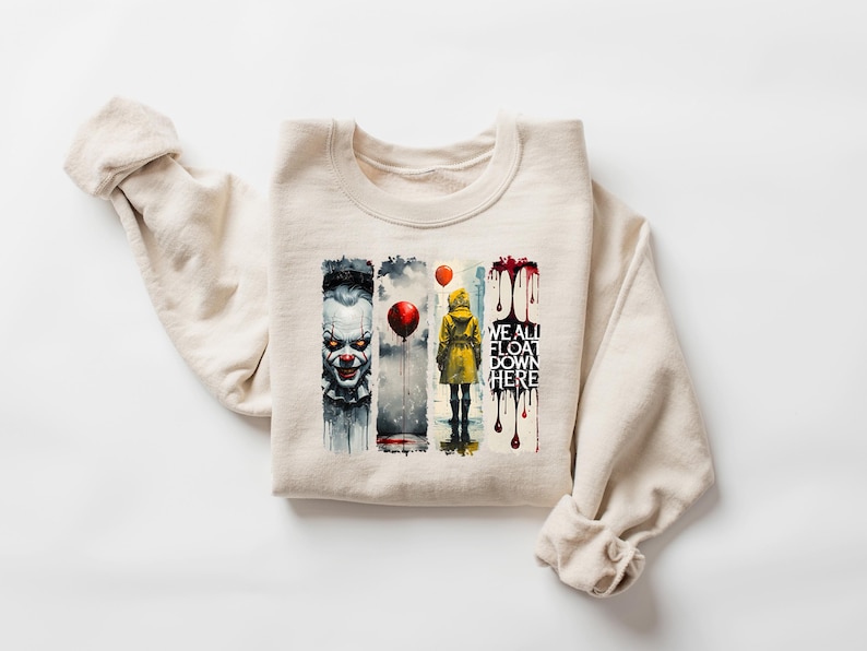 May include: Beige crewneck sweatshirt featuring a watercolor design with a scary clown, a person in a yellow raincoat holding a red balloon, and the text "WE ALL FLOAT DOWN HERE".