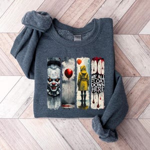 May include: Dark gray sweatshirt featuring a graphic design with a scary clown, a person in a yellow raincoat, and the text "WE ALL FLOAT DOWN HERE". The design includes red balloons and dripping blood.