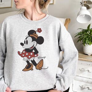 May include: A light gray sweatshirt featuring a graphic of Minnie Mouse in a classic pose. Minnie is wearing a red polka-dot skirt, brown shoes, and a matching hat with a flower. The Disney logo is printed on the bottom right.