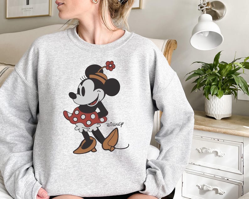 May include: A light gray sweatshirt featuring a graphic of Minnie Mouse in a classic pose. Minnie is wearing a red polka-dot skirt, brown shoes, and a matching hat with a flower. The Disney logo is printed on the bottom right.