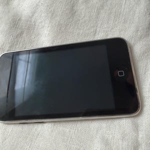 May include: A rectangular digital media player with a black screen and a silver metallic frame. The device features a single square home button centered below the display screen, which is positioned horizontally on a textured off-white surface.