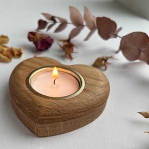 May include: A heart-shaped wooden candle holder with a lit white tea light candle. The candle holder has a gold-coloured metal rim. Dried flowers and leaves are scattered around the candle holder on a white surface.