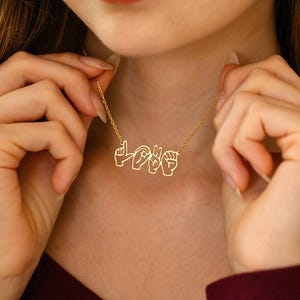 Puede incluir: Un collar dorado que deletrea "LOVE" en lenguaje de señas americano. El colgante es un diseño delicado de alambre. El collar se lleva alrededor del cuello, con las manos de una persona sosteniendo la cadena.
