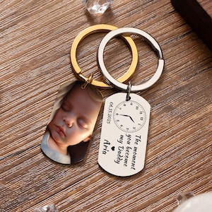 Op de afbeelding: Een gepersonaliseerde sleutelhanger met een foto van een slapende baby en een label met de tekst "The moment you become my Daddy" en de naam "Aria". De sleutelhanger heeft gouden en zilveren ringen.