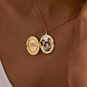 May include: An oval gold locket necklace on a thin chain. The locket is open, revealing a photograph of three people on the right side and the engraved text Always with me on the left side. The metal has a polished finish and a classic hinge design.