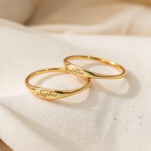 May include: Two polished gold-tone rings featuring a smooth, rounded band design. Each ring is engraved with a name in a cursive script font, with one ring displaying the name Sophia and the other ring displaying the name Alex.
