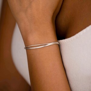 Puede incluir: Una pulsera de plata delicada que presenta dos bandas paralelas, finas y lisas, con una ligera curva. El accesorio metálico tiene un acabado pulido y descansa sobre la muñeca, mostrando un diseño simple y limpio con una forma circular continua.