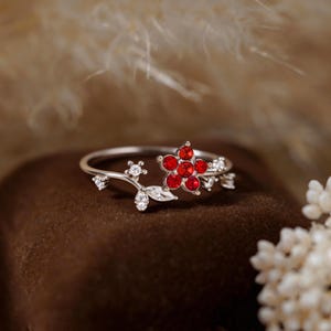 May include: A silver-toned ring featuring a delicate vine-like band adorned with a cluster of five vibrant red circular stones forming a flower shape, accented by several small clear white stones shaped like leaves and buds along the metal stem.