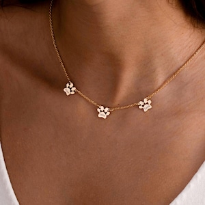 Puede incluir: Un collar de tono dorado que presenta tres pequeños dijes con forma de huella de animal espaciados a lo largo de una cadena delicada. Cada dije está grabado con un nombre, incluyendo Ollie y Luna, creando una pieza de joyería personalizada.