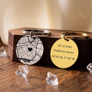 May include: Two personalised keyrings. One has a black and white map design with a heart, the other is gold with engraved text: "30.12.2025 Sophia & James 38°53' N, 77°02' W". Both have metal key rings and are on a wooden box.
