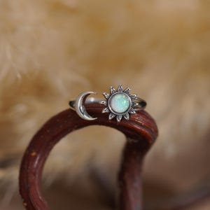 May include: A silver-toned open ring featuring a crescent moon on one end and a sun motif on the other. The sun design is centered with a round, iridescent white stone that displays flecks of color. The metal has a darkened, oxidized finish.