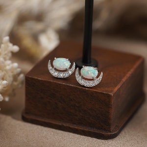 May include: A pair of stud earrings featuring an oval-shaped iridescent opal stone set above a silver-toned crescent moon shape. The crescent moon is embellished with small, clear, sparkling stones along its curved surface.