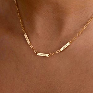 Può includere: Una collana a catena color oro con tre piccole barre metalliche rettangolari integrate nelle maglie. Ogni barra è incisa con un nome: Ezra, Mason e Chloe. La collana presenta un design a maglie sottili e una finitura metallica lucida.