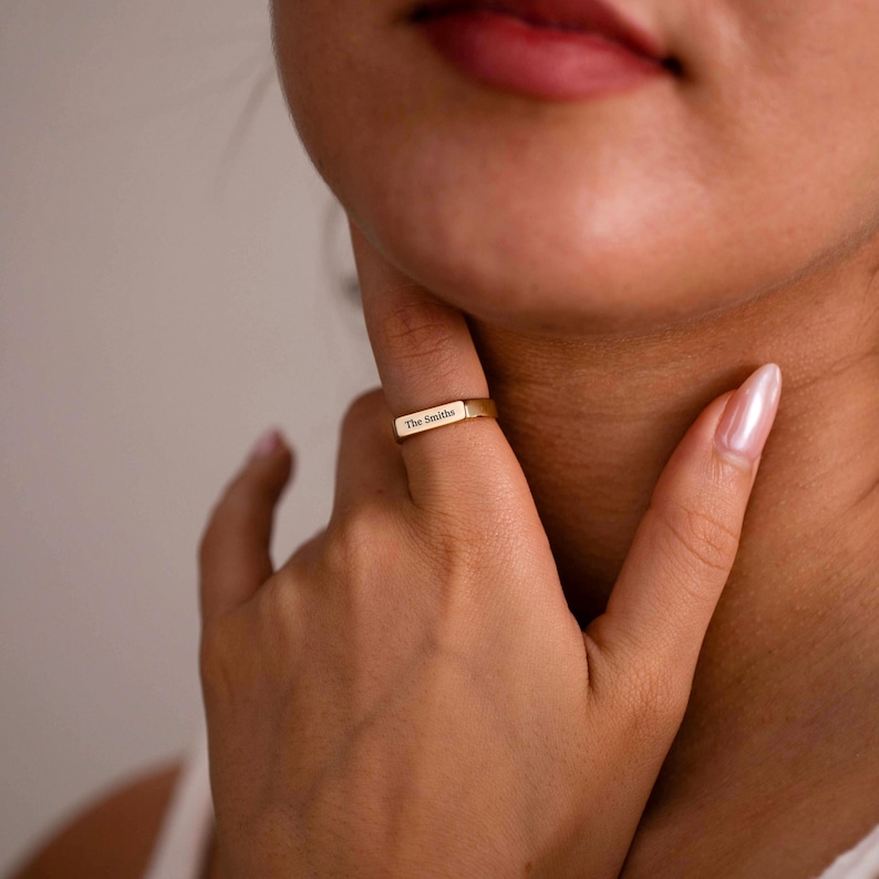 May include: A gold-toned metal ring featuring a flat, rectangular bar surface. The bar is engraved with the text "The Smiths" in a clear, dark font. The ring has a smooth, polished finish and a simple, clean design that sits horizontally on the finger.