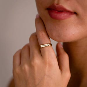 May include: A gold-toned metal ring featuring a flat, rectangular bar surface. The bar is engraved with the date 02.05.2025 in a clear, dark font. The ring has a smooth, polished finish and a slim band that encircles the finger.