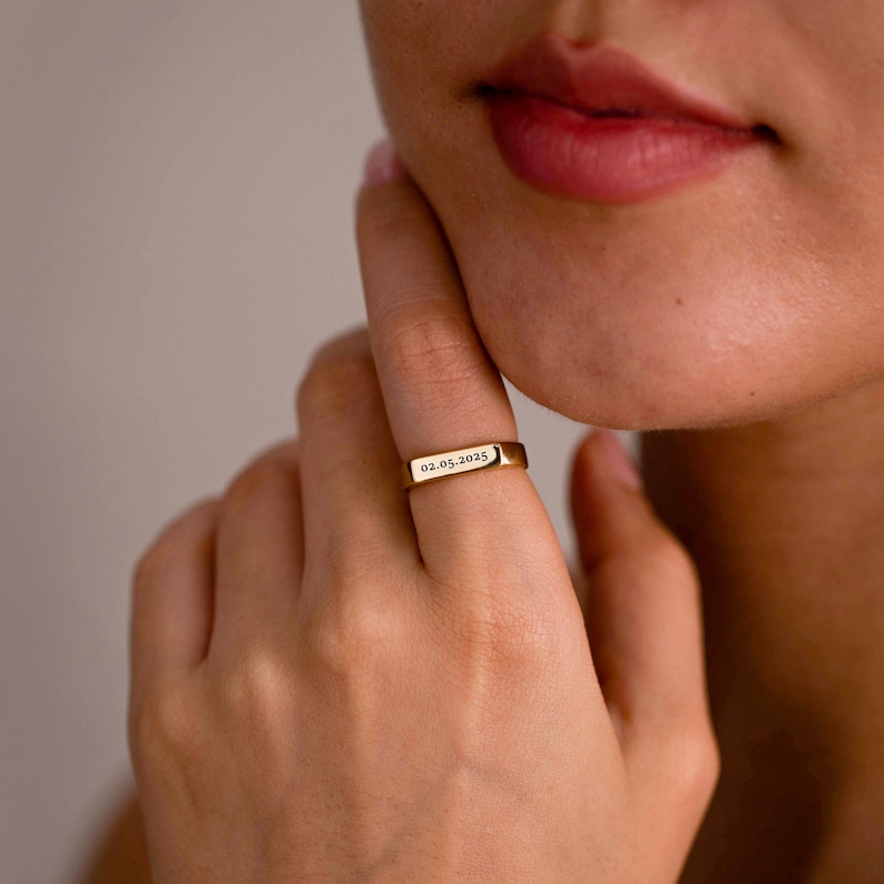 May include: A gold-toned metal ring featuring a flat, rectangular bar surface. The bar is engraved with the date 02.05.2025 in a clear, dark font. The ring has a smooth, polished finish and a slim band that encircles the finger.
