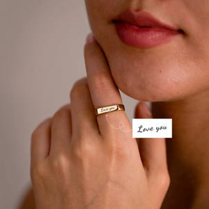 May include: A gold-toned band ring featuring a flat, rectangular front surface engraved with the cursive text "Love you". The ring has a smooth, polished finish and is positioned on a finger, with a callout box repeating the text "Love you".