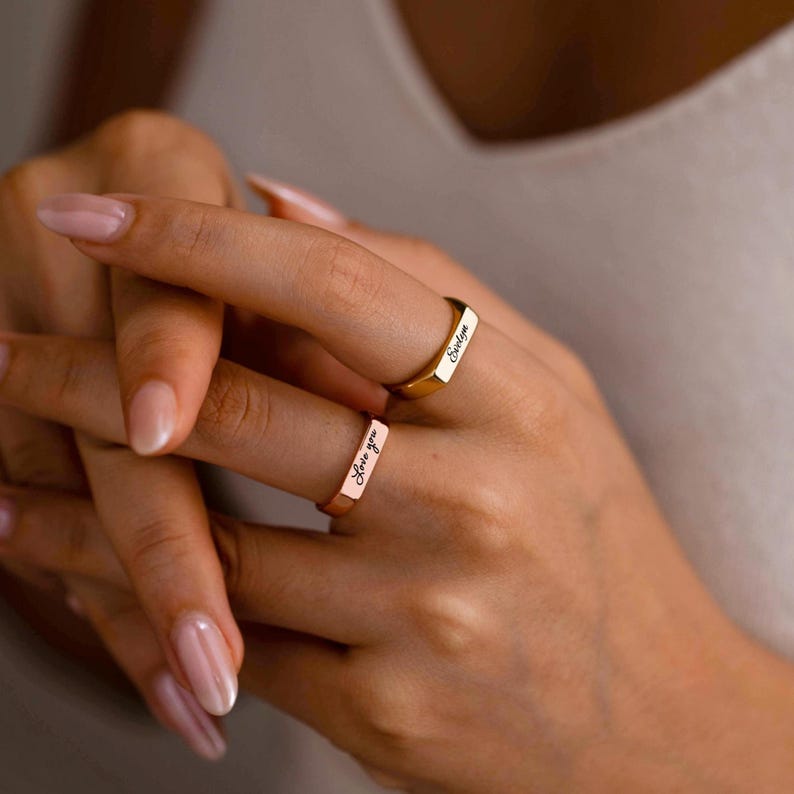 May include: Two metal rings with flat rectangular surfaces. One ring is gold-toned and engraved with the name Evelyn in a cursive script. The second ring is rose gold-toned and engraved with the phrase Love you in a matching cursive script.