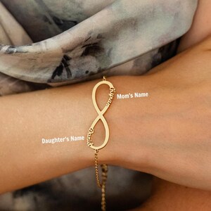 Peut inclure: Un bracelet doré avec un pendentif central en forme de symbole infini. Le pendentif est gravé avec les noms Kyran et Danik le long de ses bords incurvés. Le bracelet est fixé à une fine chaîne dorée qui s'attache autour du poignet.