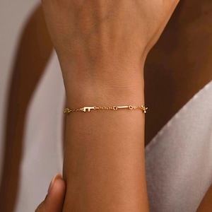 Op de afbeelding: Een delicate gouden kettingarmband met een kleine, gestileerde letter F-bedel geïntegreerd in de schakels. De armband heeft een gepolijste afwerking en een dun, verfijnd ontwerp dat horizontaal over de pols rust, met een eenvoudige schakelstructuur.