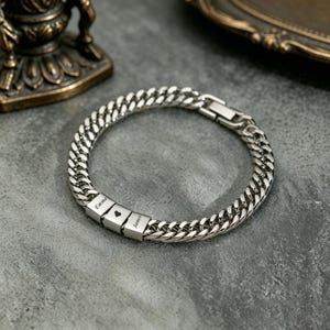 Puede incluir: Una pulsera de cadena de bordillo plateada con tres cuentas cuadradas. Las cuentas están grabadas con los nombres "Emma" y "James", y un símbolo de corazón. La pulsera está sobre una superficie gris.