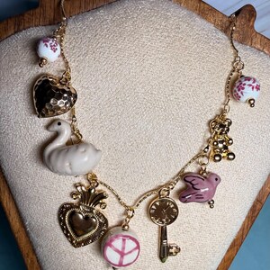 May include: A gold chain necklace with various charms, including a heart, a swan, a dove, a peace sign, a key, and a clock. The charms are made of ceramic and metal.