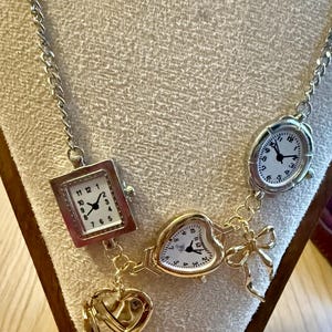 May include: Silver-toned necklace featuring multiple miniature watch faces in square, oval, and heart shapes. The necklace includes a heart-shaped charm and a bow charm. The watch faces have white backgrounds with black numbers and hands.