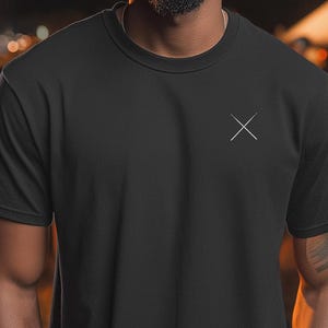 Puede incluir: Camiseta negra de cuello redondo con un pequeño diseño de cruz blanca en el pecho izquierdo. La camiseta de manga corta está hecha de un material suave. El fondo está borroso con luces y personas.