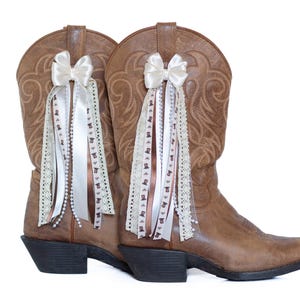 May include: A pair of brown leather cowboy boots with decorative bows and ribbons. The bows are white, and the ribbons are white, brown, and cream-colored with lace and pearl accents. The boots have a classic western design.