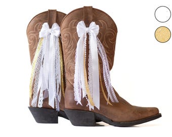 May include: A pair of brown leather cowboy boots adorned with white satin bows and flowing ribbons. The ribbons include white lace, gold glitter, and white satin, adding a touch of elegance to the Western-style footwear.