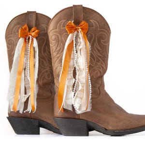 May include: Two brown leather cowboy boots, each decorated with an orange bow and flowing ribbons. The boots feature detailed stitching and are embellished with white beaded strands. The bows and ribbons add a rustic charm.