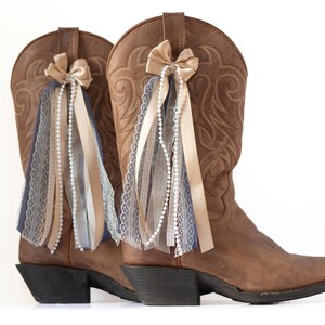 May include: A pair of brown leather cowboy boots with decorative bows and ribbons. The boots are adorned with cream-colored bows, pearl strands, and ribbons in cream, blue, and lace. The boots have a classic western design.